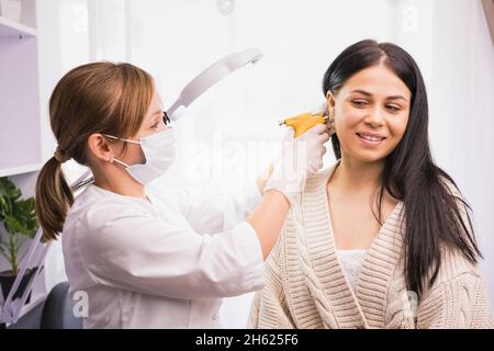 Un spécialiste du masque médical et des gants piquent les oreilles du client à l'aide d'un pistolet de perçage dans l'armoire du cosmetologiste. Banque D'Images