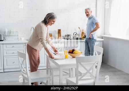 Femme souriante mettant des assiettes sur la table près de jus d'orange, fruits et mari dans la cuisine Banque D'Images