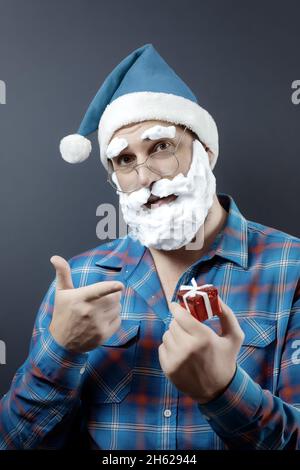 Un homme dans un chapeau de père noël et une barbe en mousse tient une boîte cadeau rouge minuscule et pointe son index vers elle Banque D'Images