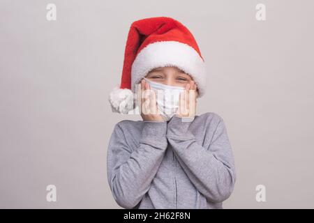 Un garçon dans un chapeau d'aide de Santa et un masque de protection médicale tient un masque avec ses mains. Banque D'Images