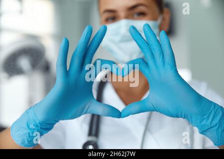 Femme avec un masque médical et les mains dans le gant de latex montre le symbole du coeur.Médecin pour le cœur.Aimez nos professionnels de la santé. Banque D'Images