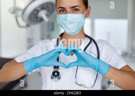 Femme avec un masque médical et les mains dans le gant de latex montre le symbole du coeur.Médecin pour le cœur.Aimez nos professionnels de la santé. Banque D'Images