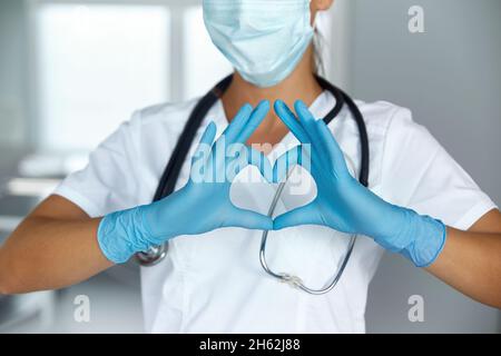 Femme avec un masque médical et les mains dans le gant de latex montre le symbole du coeur.Médecin pour le cœur.Aimez nos professionnels de la santé. Banque D'Images