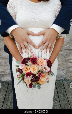 femme enceinte en robe de dentelle blanche avec le bouquet de mariée coloré, le mari tient le ventre de grossesse Banque D'Images