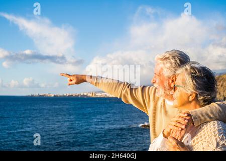 portrait de deux personnes âgées et matures appréciant l'été sur la plage en regardant la mer souriant et en s'amusant avec le coucher du soleil à l'arrière-plan. deux personnes âgées actives voyageant à l'extérieur. Banque D'Images