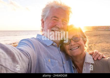 portrait de couple de personnes âgées et matures profitant de l'été à la plage en regardant l'appareil photo prendre un selfie avec le coucher du soleil à l'arrière-plan. deux aînés actifs voyageant à l'extérieur. Banque D'Images