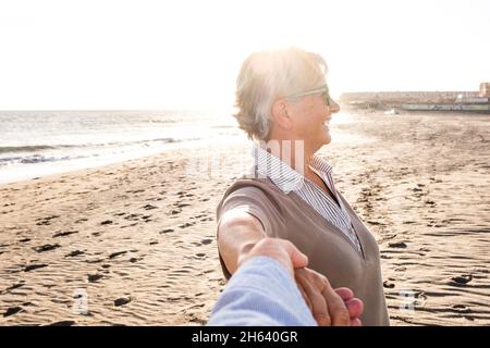 première vue et point de vue de l'homme mûr et vieux tenant la main de sa femme à la plage, s'amuser et profiter ensemble l'été. deux aînés heureux dehors avec le coucher du soleil à l'arrière-plan. Banque D'Images