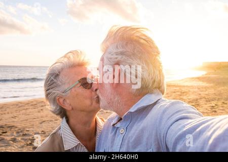 portrait de couple de personnes âgées et matures profitant de l'été à la plage en regardant l'appareil photo prendre un selfie avec le coucher du soleil à l'arrière-plan. deux aînés actifs voyageant à l'extérieur. Banque D'Images