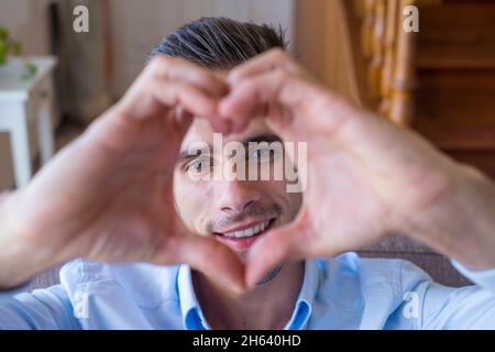 un adolescent affectueux et joyeux prend un portrait romantique de selfie en regardant l'appareil photo à travers le coeur des doigts joints. un jeune garçon épris montrant son amour à l'appareil photo faisant une forme de coeur avec ses mains Banque D'Images