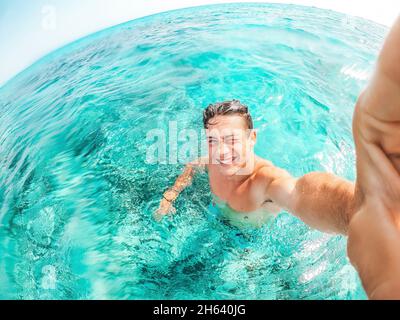 portrait d'un jeune homme heureux prenant un selfie de lui sur la plage dans une eau turquoise bleue ayant du plaisir et appréciant seul des vacances à l'extérieur. Banque D'Images