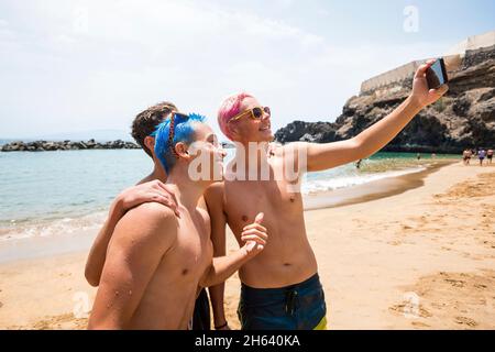 groupe de trois adolescents heureux et gaies, les garçons appréciant et souriant regardant l'appareil photo prendre un selfie ensemble à la plage. Banque D'Images