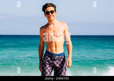 jeune adolescent beau en maillot de bain marchant sur la plage avec le ciel bleu et l'eau de l'océan en arrière-plan appréciant l'été vacances en plein air souriant et regardant sur la caméra Banque D'Images