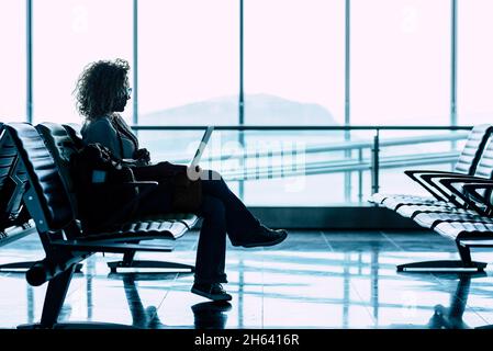 une femme s'assoit seule à la porte de l'aéroport en attendant le vol pour voyager - voyage d'affaires ou de vacances - fenêtre lumineuse en arrière-plan - femme assise et attente retard départ avion Banque D'Images