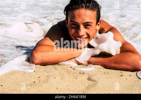 joyeux et gai jeune caucasien beau garçon se posant sur la plage et de jouer avec vague mousse smiling et rire dans les vacances d'été style de vie Banque D'Images