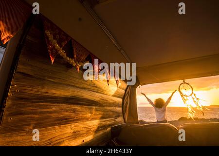 vue arrière d'une femme aux bras étendus, vue panoramique sur les paysages marins au coucher du soleil à côté de dreamcatcher suspendu de la camionnette. femme aux cheveux bouclés appréciant ses vacances à la plage par dreamcatcher Banque D'Images