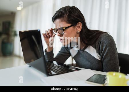 Femme latine avec des problèmes de vision essayant de lire du contenu en ligne sur un ordinateur portable à la maison. Banque D'Images