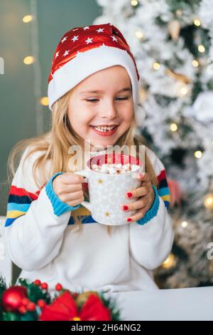 Petite fille souriante Santa aide boire du chocolat chaud avec des guimauves de tasse de noël avec arbre de noël sur fond. Banque D'Images