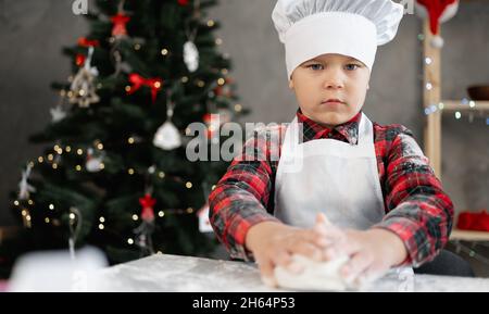 Portrait d'un petit boulanger en uniforme faisant de la pâte à pain.Garçon prépare des biscuits ou du pain d'épice pour Noël.L'enfant fait de la pâte à pizza. Banque D'Images