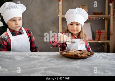 Les enfants cuisent du pain d'épice de Noël.Frère et sœur décorent les biscuits avec du sucre en poudre dans la cuisine.Petite fille boulangère.Gâteaux faits maison. Banque D'Images