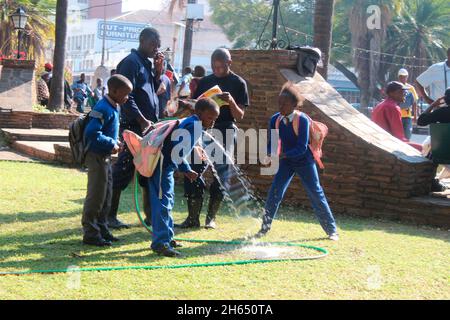 Les enfants sont vus boire de l'eau à partir d'un hospe sur la place de l'unité africaine, tandis que le gardien du parc lit l'un de leurs livres.Harare, Zimbabwe. Banque D'Images