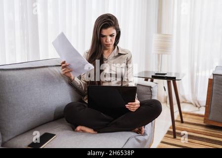 Jeune femme brésilienne belle travaillant avec un ordinateur portable tout en étant assis au canapé, travaillant à la maison concept. Banque D'Images