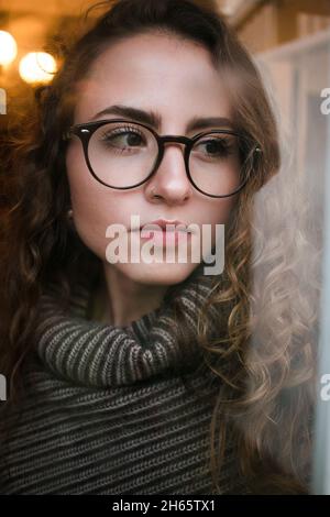 Portrait chaud et hivernal de jeune adulte, femme, regardant par la fenêtre avec des gouttes d'eau et de bokeh Banque D'Images