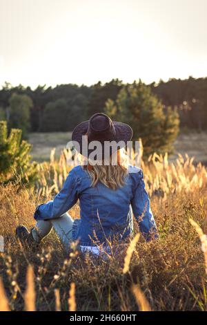 Femme assise dans l'herbe et profitant du coucher de soleil dans la nature.Détente à l'extérieur.Femme portant une veste en denim et un chapeau de cow-boy Banque D'Images