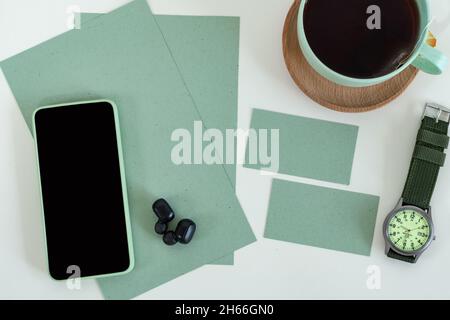 Vue de dessus d'un smartphone, d'écouteurs, d'une carte de visite, d'une tasse à thé et de montres sur un bureau vert moderne Banque D'Images