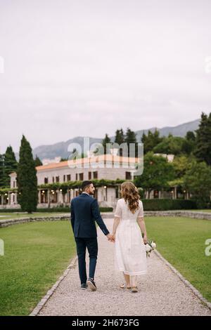 Marié et mariée dans une robe blanche, tenant les mains, marchez le long du chemin entre le jardin à la vieille villa parmi les arbres verts Banque D'Images