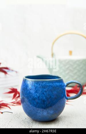 Mug bleu clair avec théière verte, feuilles d'érable rouge et livres sur fond blanc.Espace de texte. Banque D'Images