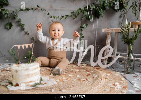 Joyeux anniversaire un an tout-petit, garçon avec gâteau assis sur le sol, fond naturel, décor de style rustique. Banque D'Images