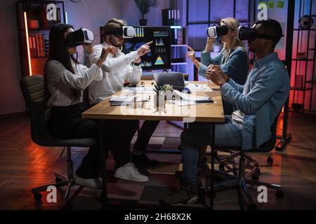 Groupe de quatre collègues multiraciaux portant un casque VR lors d'une réunion de travail au bureau.Partenaires d'affaires assis au bureau et touchant l'écran virtuel. Banque D'Images