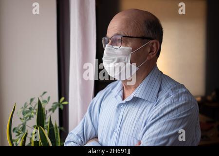 Homme âgé de race blanche portant un masque facial regardant dans la rue près de la fenêtre Banque D'Images