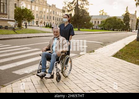 Jeune infirmière en masque de protection aidant un homme handicapé en fauteuil roulant pendant une promenade dans la ville Banque D'Images