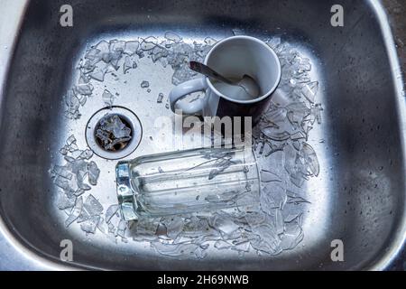 un verre brisé dans un tas de feuilles de verre et une tasse en céramique avec une cuillère se trouvent dans un évier de cuisine en acier Banque D'Images