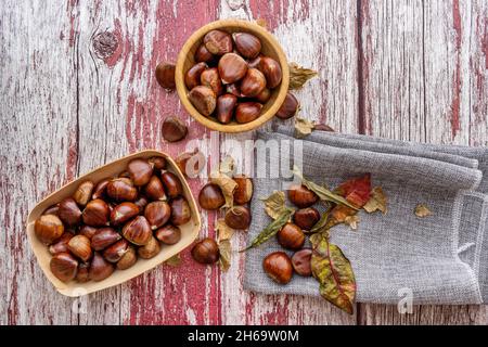 Châtaignes comestibles crues et feuilles d'automne sèches sur table en planche avec des traces de peinture rouge.Récipients en bois et serviette de cuisine grise Banque D'Images