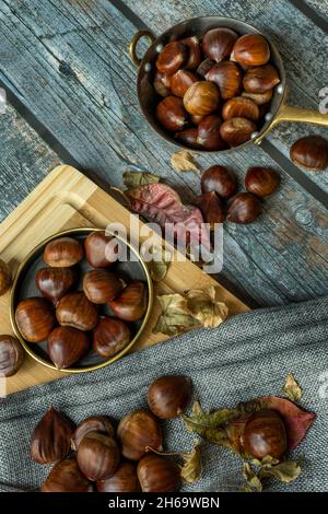 Encore la vie avec une casserole en métal pleine de châtaignes et de feuilles d'automne séchées sur la surface de la planche grise et table en bambou avec torchon Banque D'Images