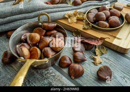 Encore la vie avec une casserole en métal pleine de châtaignes et de feuilles d'automne sèches sur la surface grise et la table en bambou Banque D'Images