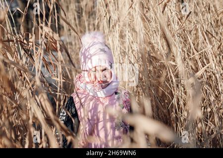 L'enfant est debout dans l'herbe sèche.Petite fille en vêtements d'hiver roses aime le soleil et explore un épais de roseaux le jour ensoleillé dans la nature Banque D'Images