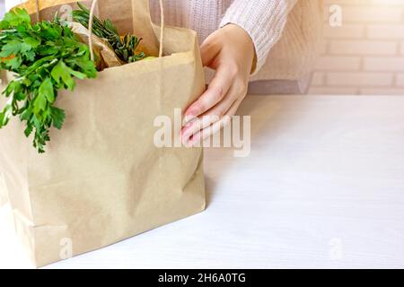 Les mains de la femme caucasienne portent un sac de papier d'artisanat avec des légumes verts de la livraison en ligne de shopping à la maison dans la cuisine sur fond clair avec la copie spac Banque D'Images
