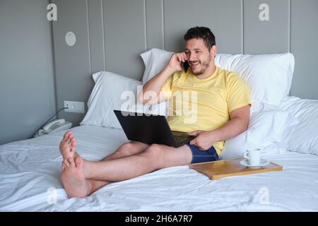 Un homme désespéré parle au téléphone et travaille avec un ordinateur portable dans un lit d'hôtel. Banque D'Images