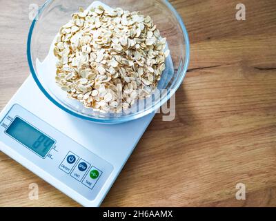 Gruaux d'avoine, gruaux pesant dans un bol en verre sur des balances électroniques de cuisine sur une table en bois. Banque D'Images