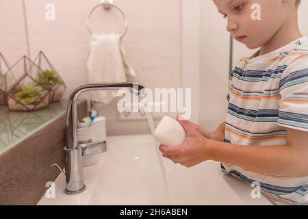 Un garçon caucasien se lave les mains dans une salle de bain légère avec du savon et de l'eau du robinet Banque D'Images