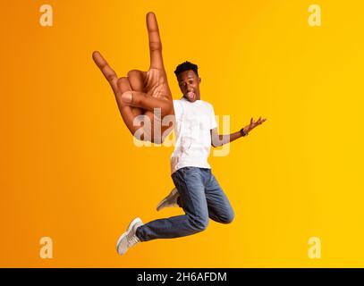 Un homme afro-américain insouciant qui saute et montre du gros rock sur le geste, un homme noir millénaire gai qui saute et gaie sur l'orange studio ba Banque D'Images