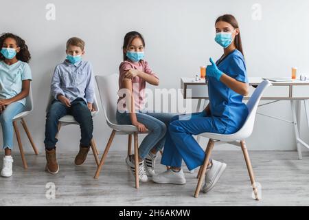 Vaccination Covid-19.Vacciné asiatique Kid fille et femme infirmière gestante Thumbs vers le haut regarder la caméra approuvant le vaccin en clinique, portant des masques de visage Banque D'Images