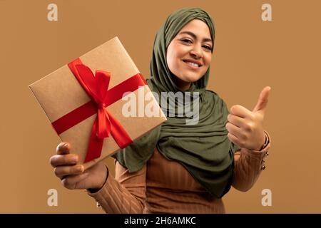 Femme musulmane tenant une boîte cadeau et montrant le pouce vers le haut, portant le hijab et souriant, posant sur fond brun de studio.Femme arabe positive tenant la naissance Banque D'Images