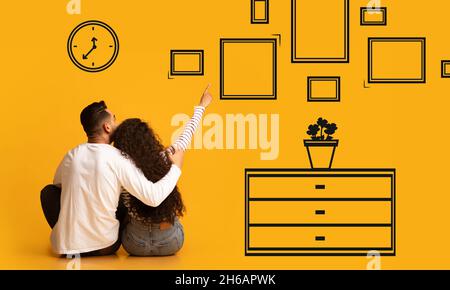 Concept d'amélioration de la maison.Arrière vue arrière du couple du millénaire pensant à la rénovation de l'appartement, les jeunes conjoints assis sur le sol près du mur jaune orange A Banque D'Images