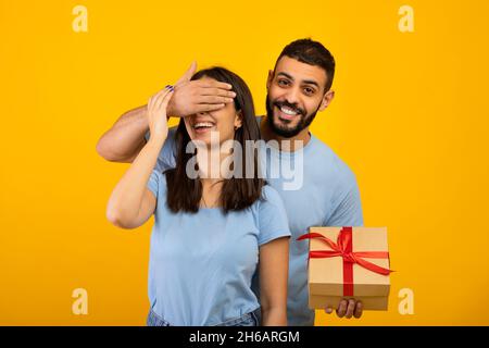 Joyeux anniversaire.Un homme arabe heureux couvrant les yeux de sa femme, saluant et donnant boîte cadeau enveloppée, debout sur fond jaune studio, Banque D'Images