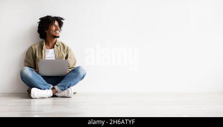 Jeune Afro-américain assis sur le sol avec un ordinateur portable et regardant de côté, souriant jeune Black Guy utilisant l'ordinateur pour le travail à distance ou l'éducation en ligne, Banque D'Images