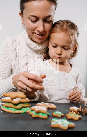 Mère avec enfant décorant des biscuits de pain d'épice avec glaçage pour Noël.Plats de fête faits maison.Atelier de famille de petite fille mignon.Étoiles, arbre Banque D'Images
